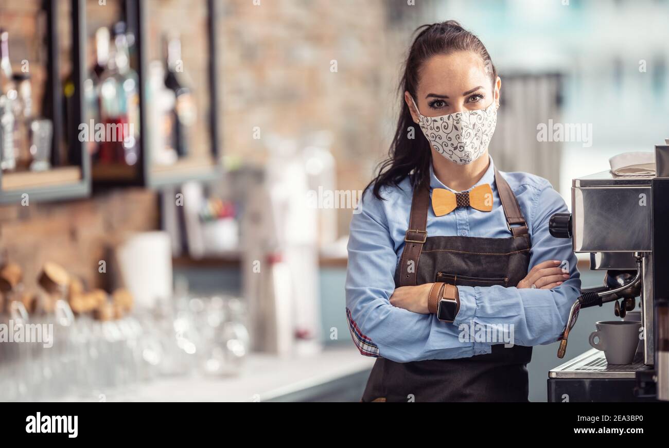 Female employee in a cafe stands unhappy with arms crossed wearing face mask. Stock Photo