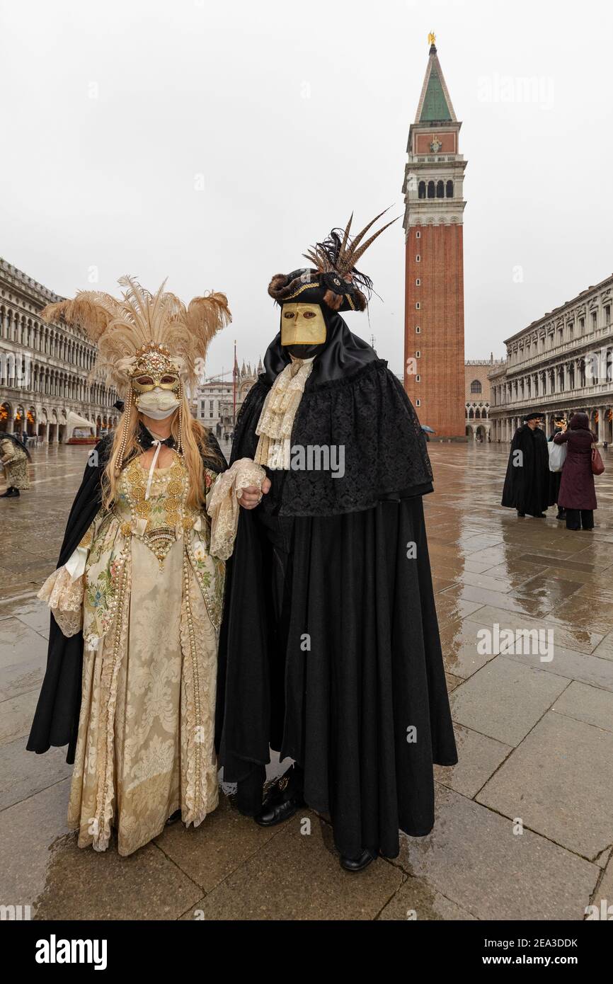 A man in a  traditional bauta costume with lava mask and feathered tricornered hat holds the hand of a lady with plumed headdress in St Mark's Square Stock Photo