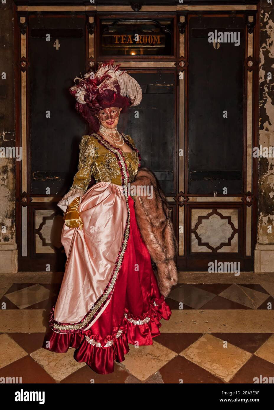 Glamourous Venetian carnival lady masked in scarlet and pink silk dress and elegant plumed hat smiling outside of traditional tea shop Stock Photo