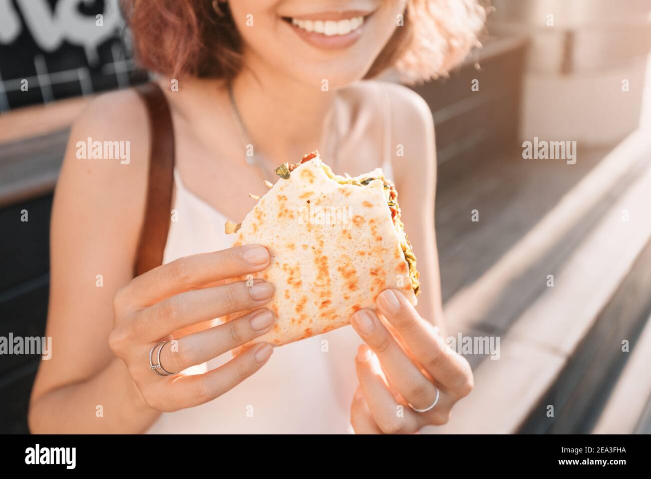 Happy woman eats a fast food flatbread with filling sitting on the street and resting after a working day. Concept of healthy food and extra calories Stock Photo