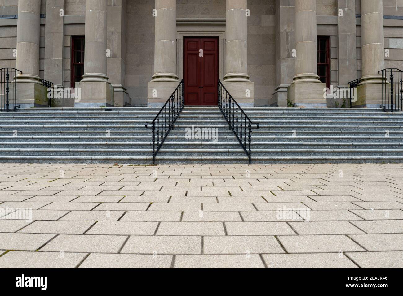Six large round concrete columns at the top of marble steps with black iron rails to a legal building. The government building has a tall red door. Stock Photo