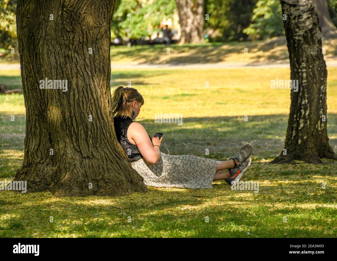 PRAGUE, CZECH REPUBLIC - JULY 2018: Person sitting down under the shade of a tree checking her phone in a public park in Prague. Stock Photo