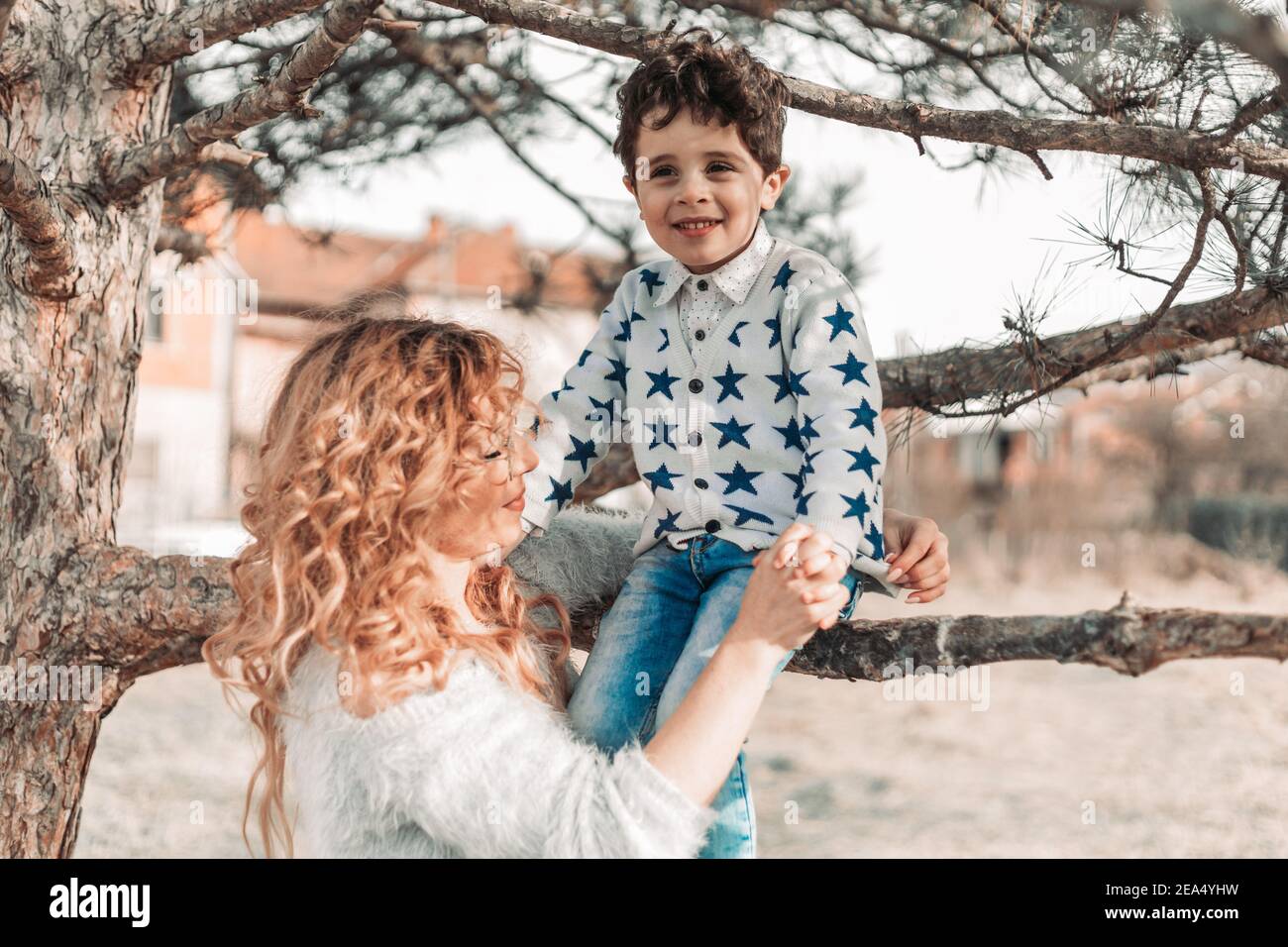 Mom holding her toddler son, while he is sitting on a tree. Love and affection Stock Photo