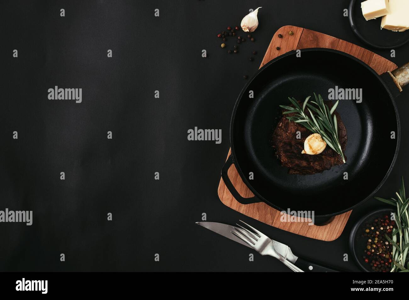 Cooked ribeye steak on castiron pan and board, rosemary, knife, fork