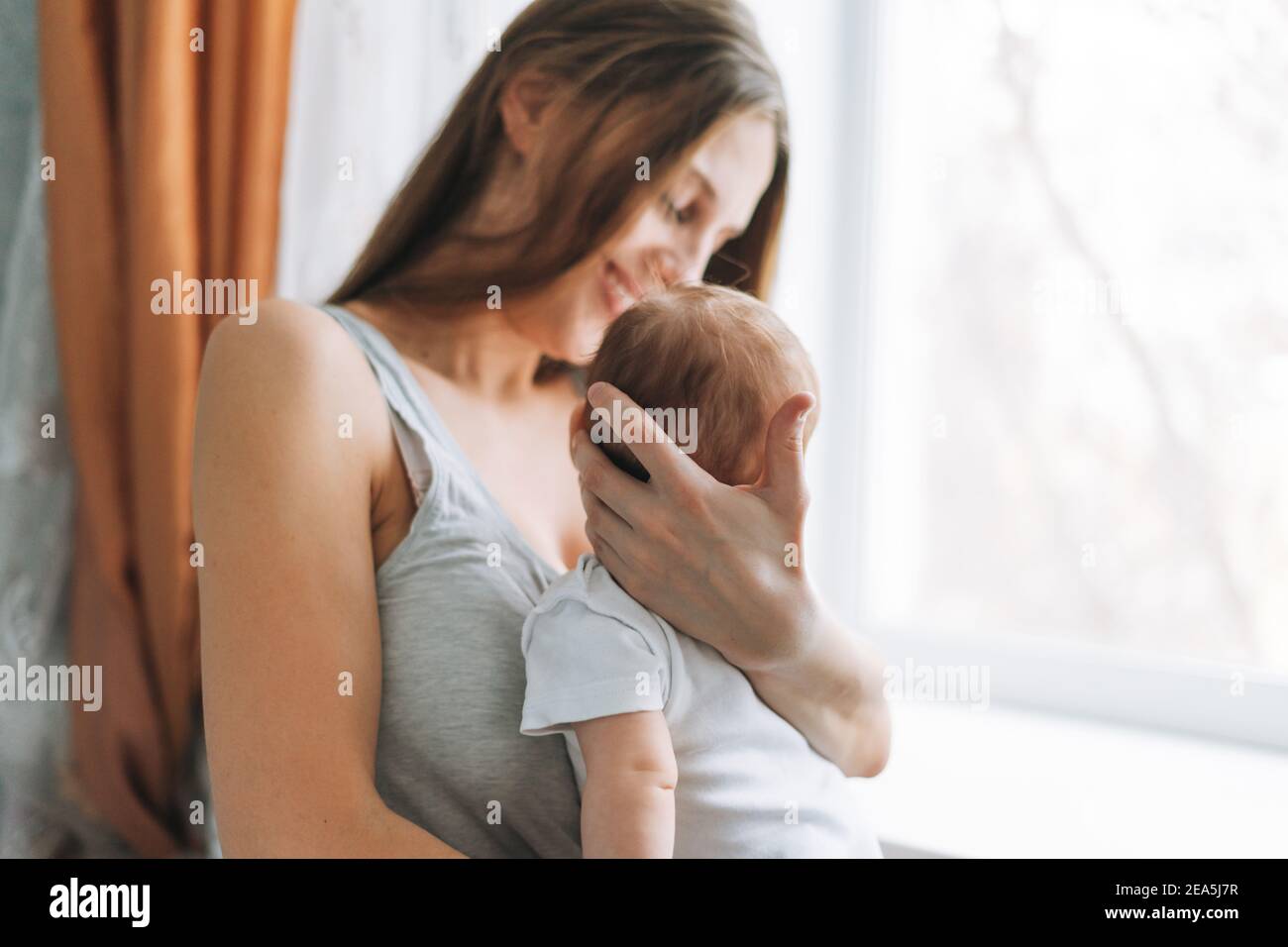 Young woman mom with long hair holding baby girl on hands near window at home Stock Photo