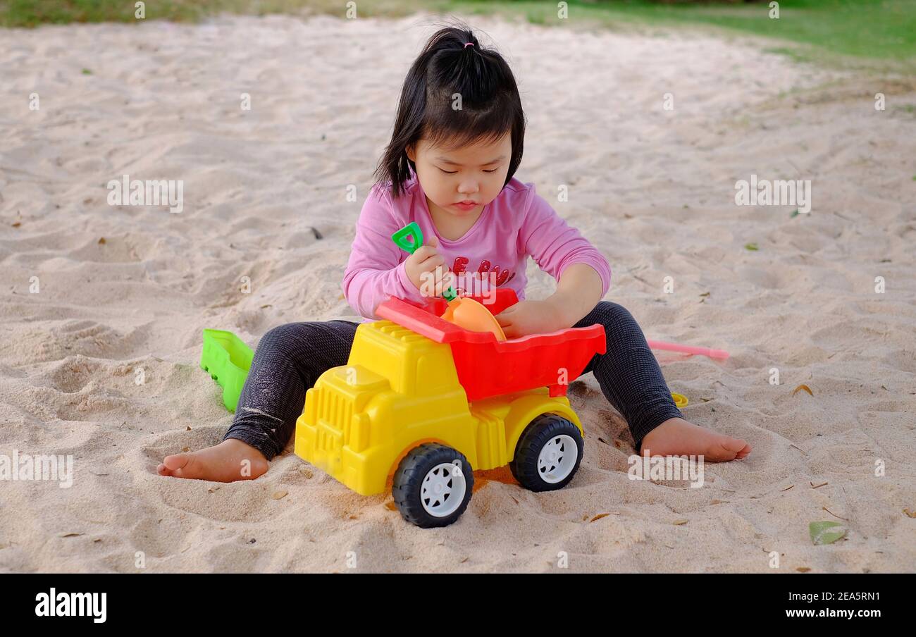 A cute young Asian girl playing with colorful plastic toys and sand in a sandbox, having fun, smiling. Stock Photo