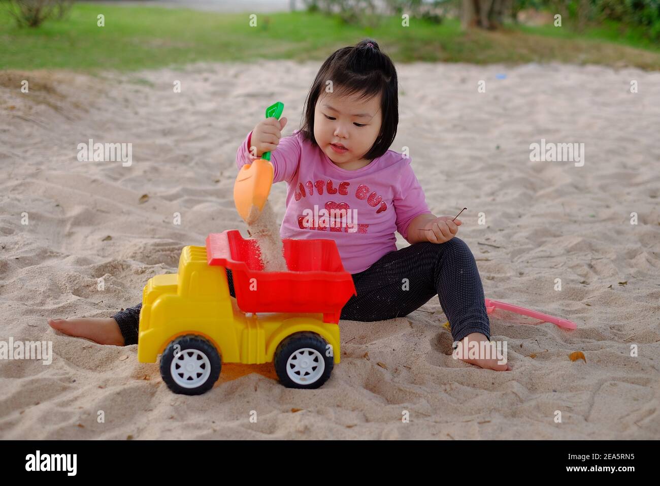 A cute young Asian girl playing with colorful plastic toys and sand in a sandbox, having fun, smiling. Stock Photo