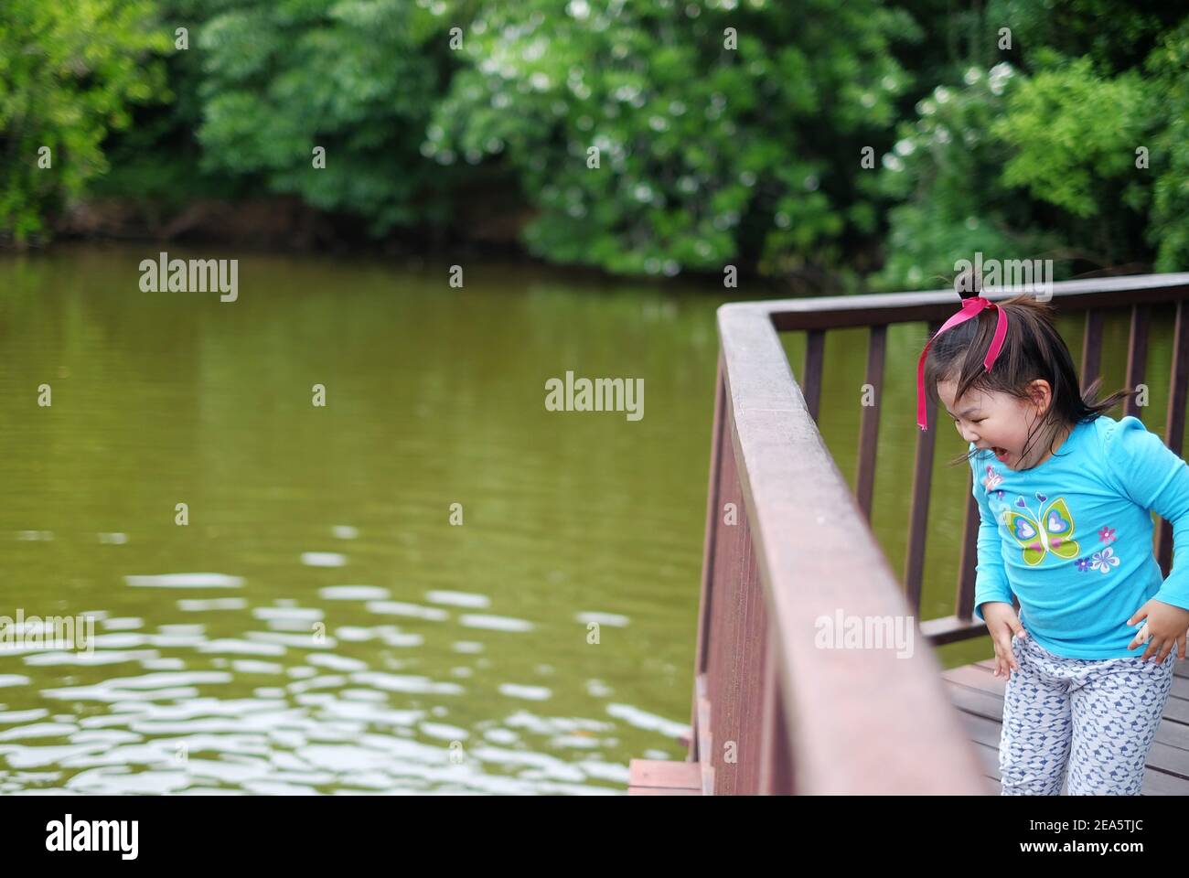 A cute young Asian girl, standing on a bridge by a pond, looking down at the water and feeling excited after seeing a turtle. Jumping up and down with Stock Photo