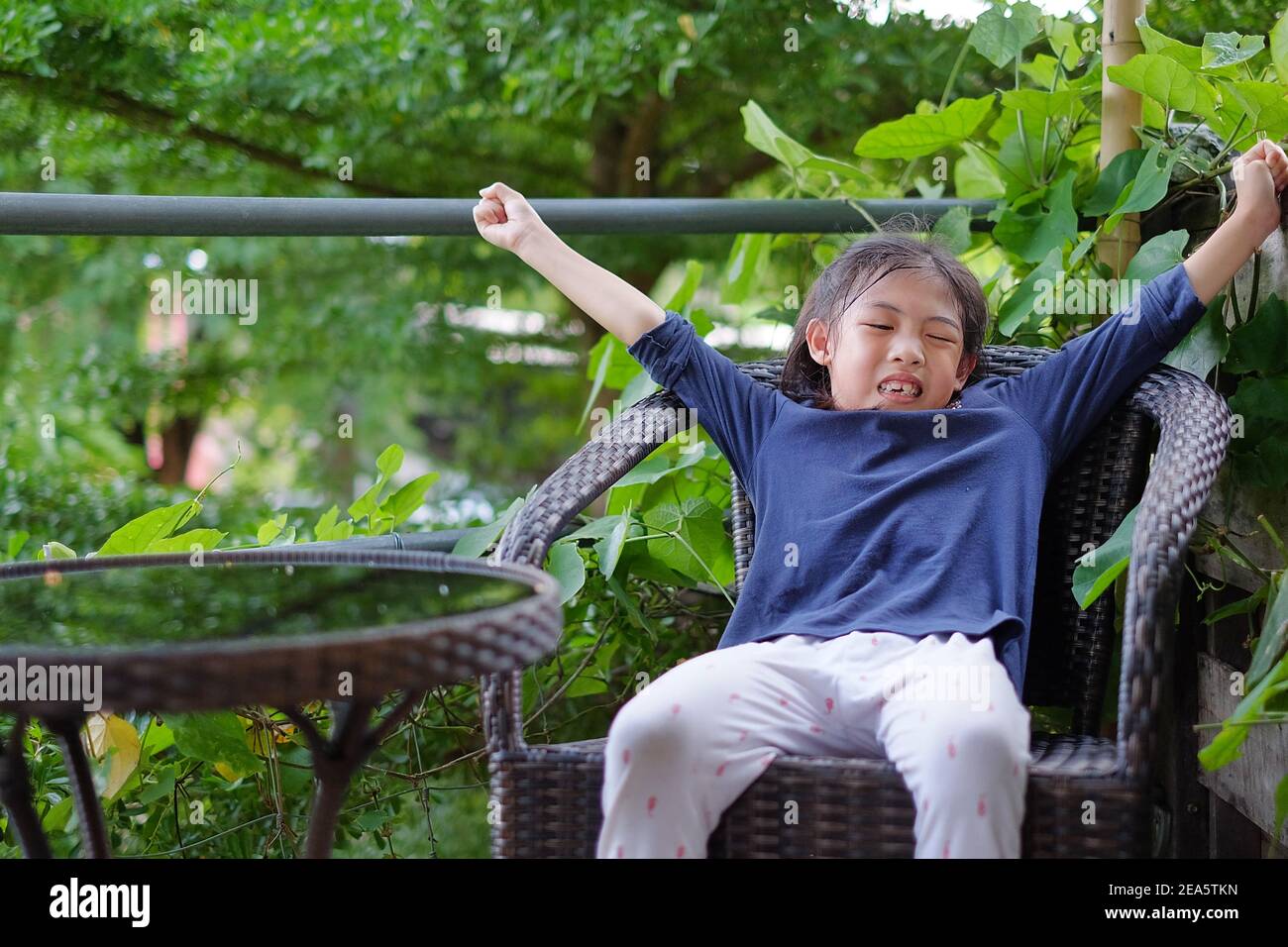 A cute young Asian girl sitting on a big ratton chair feeling bored and sleepy, stretching her arms upward with her eyes closed. Stock Photo