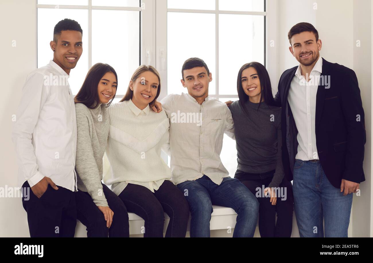Group portrait of multiracial students sitting on the windowsill and hugging looking at the camera. Stock Photo