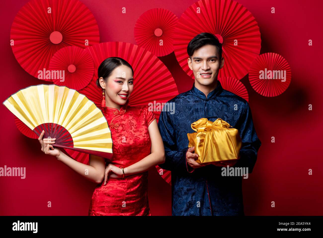 Smiling Asian couple in traditional costumes with fan and gold package as a gift for Chinese new year in oriental style red background Stock Photo