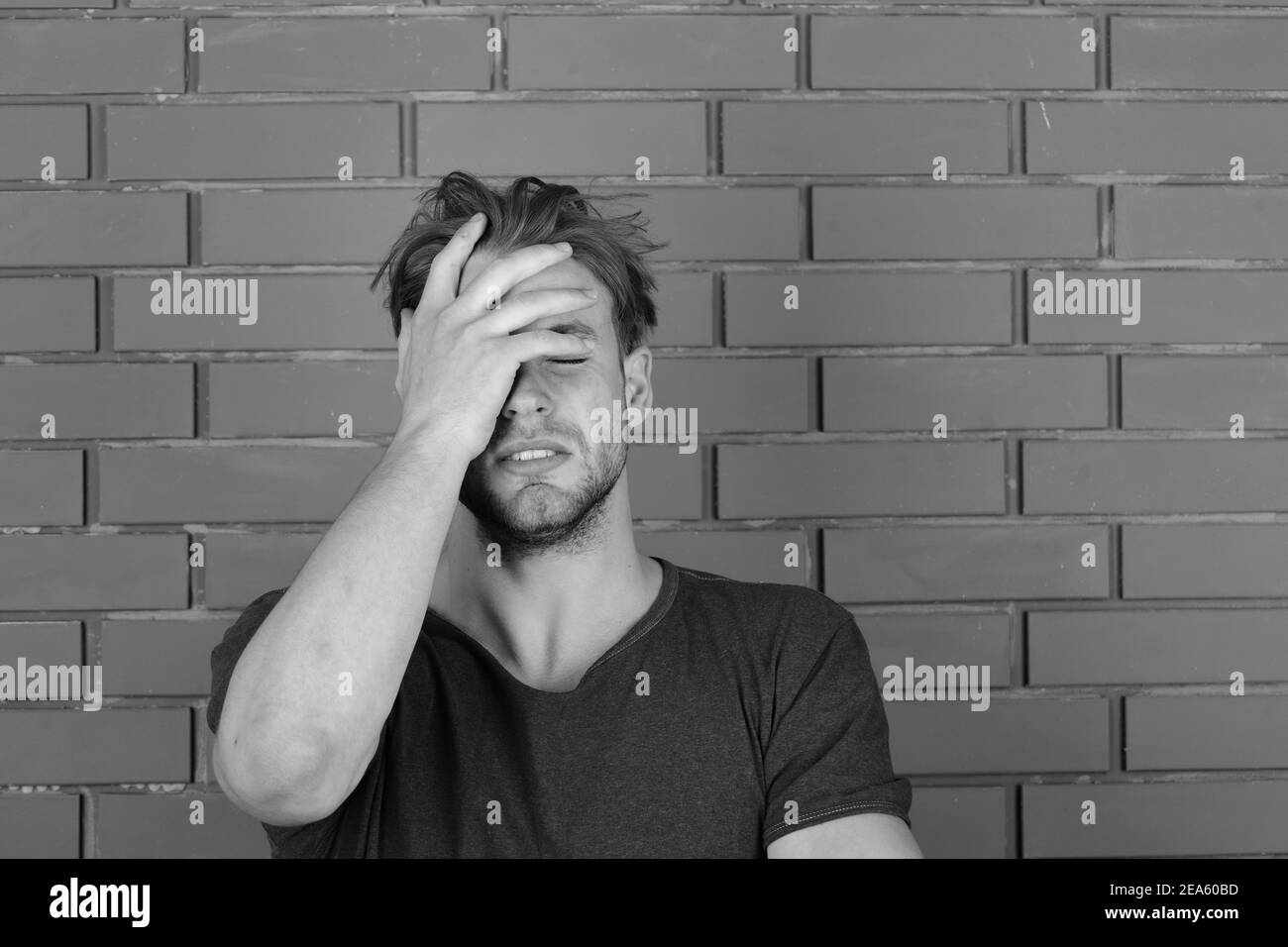 Man with fair hair on red brick wall background. Macho with suffering face holds hand on forehead. Illness and health concept. Guy with slight bristle in dark blue tshirt suffers from headache Stock Photo