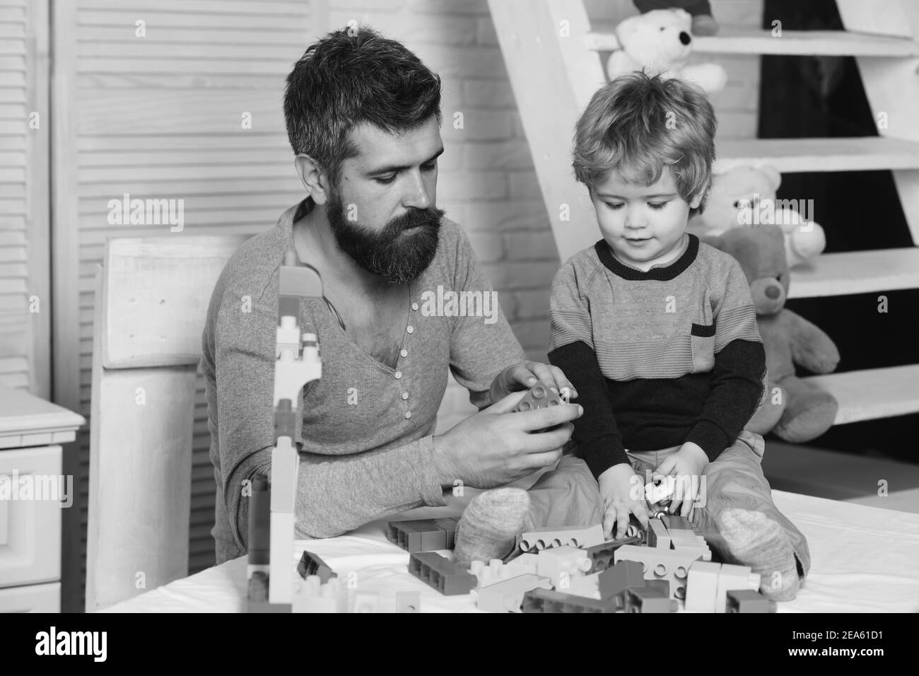 Boy and bearded man play together. Father and son with serious faces create colorful toy out of bricks. Dad and kid with toys on wooden background build of plastic blocks. Family and childhood concept Stock Photo