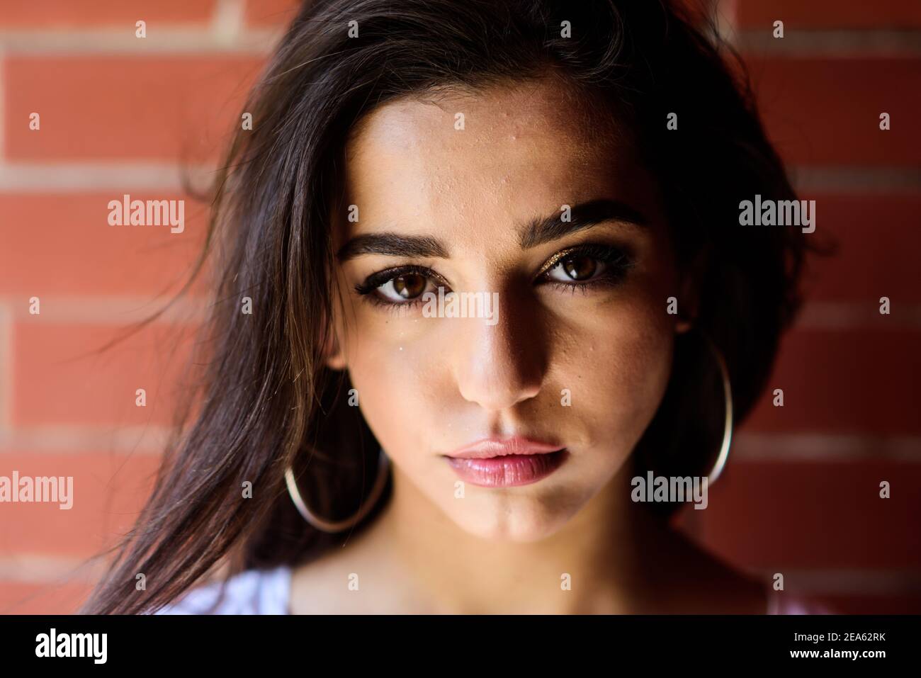 Girl attractive gorgeous brunette middle eastern appearance close up. Girl wears big metallic ring earrings accessory. Woman calm confident face with make up looks at camera. Beauty of arabian women. Stock Photo