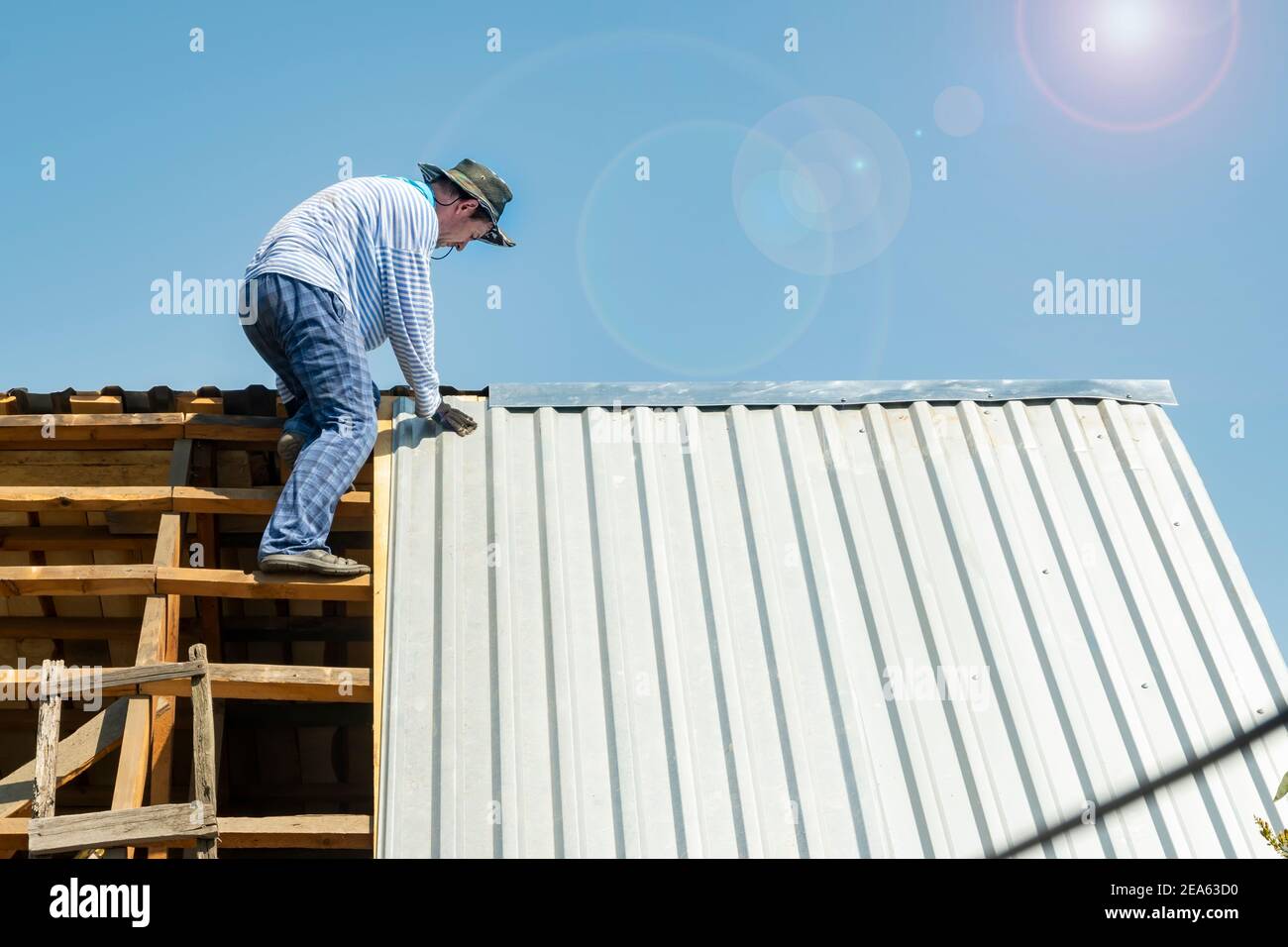 carpenter repairs the roof of the house, attaches metal galvanized sheets in hot weather in the bright sun Stock Photo