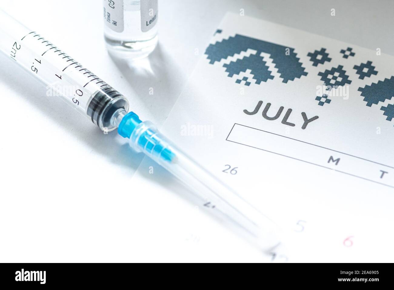 Syringe, glass vial or phial and calendar with month of July on a white table ready to be used. Covid or Coronavirus vaccine background, close up Stock Photo