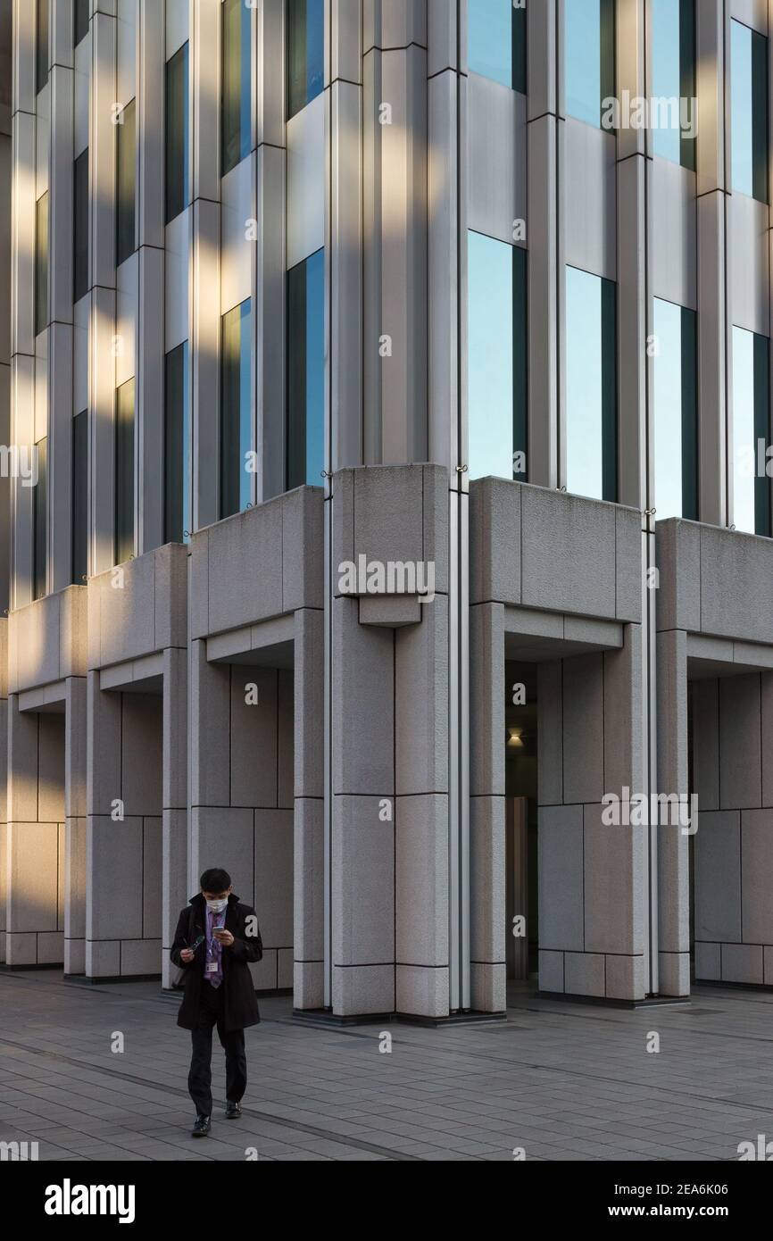 A Japanese office worker or salaryman checks his smart phone as he walks out of the Shinjuku Nomura building in Shinjuku, Tokyo, Japan Stock Photo