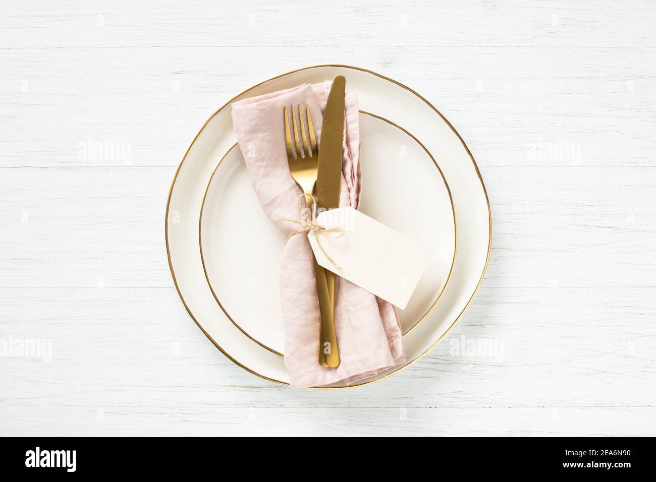 Golden rim plates and golden cutlery on pink napkin with tag on white table. Top view. Stock Photo