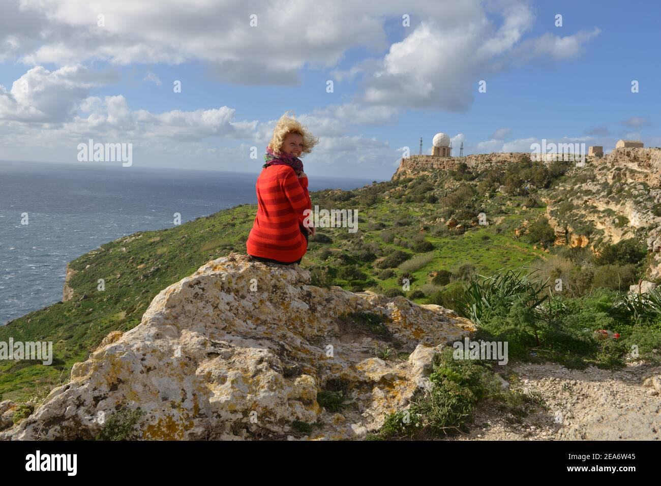 Rear view of a woman sitting on a rock looking at Dingli cliffs with Dingli Radar Station in the distance, Malta Stock Photo