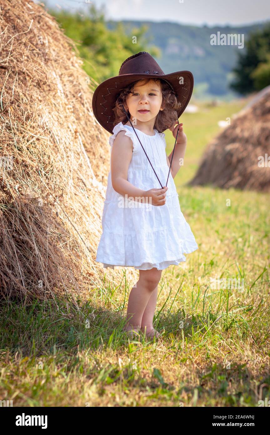 Girl wearing a cowboy hat standing in a field by a hay bale, Bulgaria Stock Photo