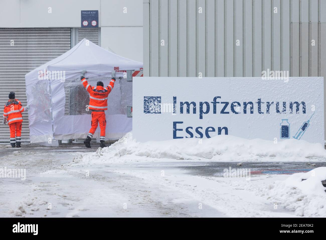 08 February 2021, North Rhine-Westphalia, Essen: Employees of 'Die Johanniter' set up a tent behind a snowy sign in front of the Essen vaccination centre. In North Rhine-Westphalia, all 53 vaccination centres start operations on Monday. First to be vaccinated are people living at home over the age of 80 who have previously made a fixed appointment. Photo: Rolf Vennenbernd/dpa Stock Photo