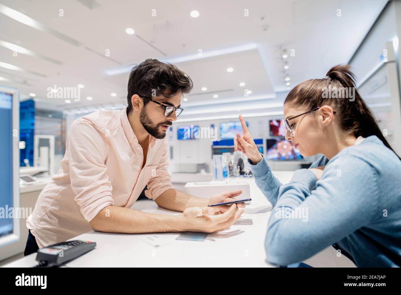 Man holding a dysfunctional smartphone while leaning on a stand. Cashier find a solution. Tech store interior. Stock Photo