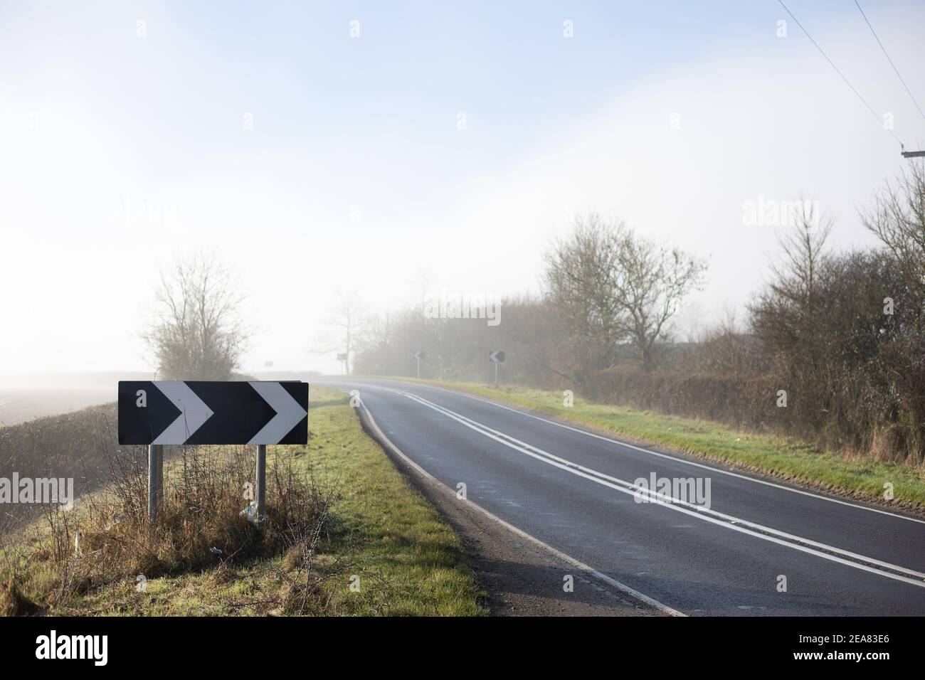 Fog covers an empty road and surrounding fields, a sign indicates it is a sharp bend. Stock Photo