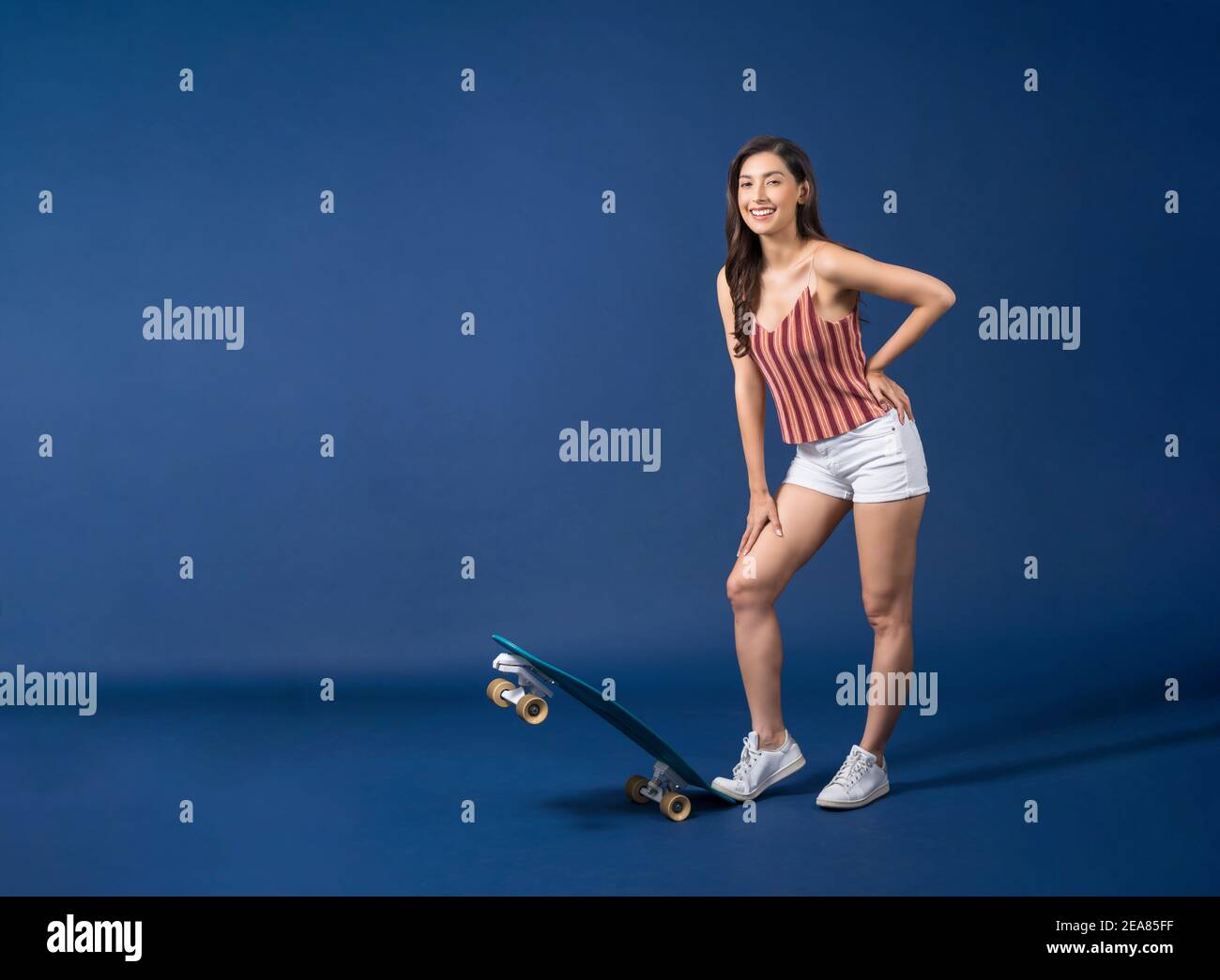 Happy young Asian woman standing on surfskate or skateboard on blue color background, exercise and sport, trendy and extream sports concept Stock Photo