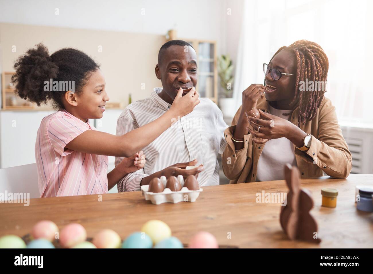 Portrait of loving African-American family making chocolate Easter decorations and tasting them while sitting at wooden table in cozy home interior Stock Photo