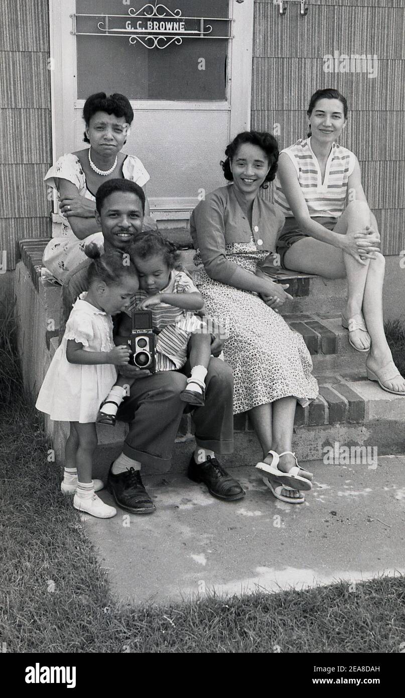 1960s, Chicago, USA, a family siiting on steps of their property, with the two youngest daughters taking an interest in their father's film camera, a Ciroflex, a twin-lens reflex camera (TLR). The Ciro Camera co started making cameras in Detroit, in 1941 and then in Delaware, Ohio, from 1947. In 1951 Graflex acquired the rights to make the camera and produced the Ciro flex twin-lens reflex.  Six models were made, the one here, the Model E, was known as the 'working man's Rolleiflex', as it was affordable for the man in the street. Stock Photo