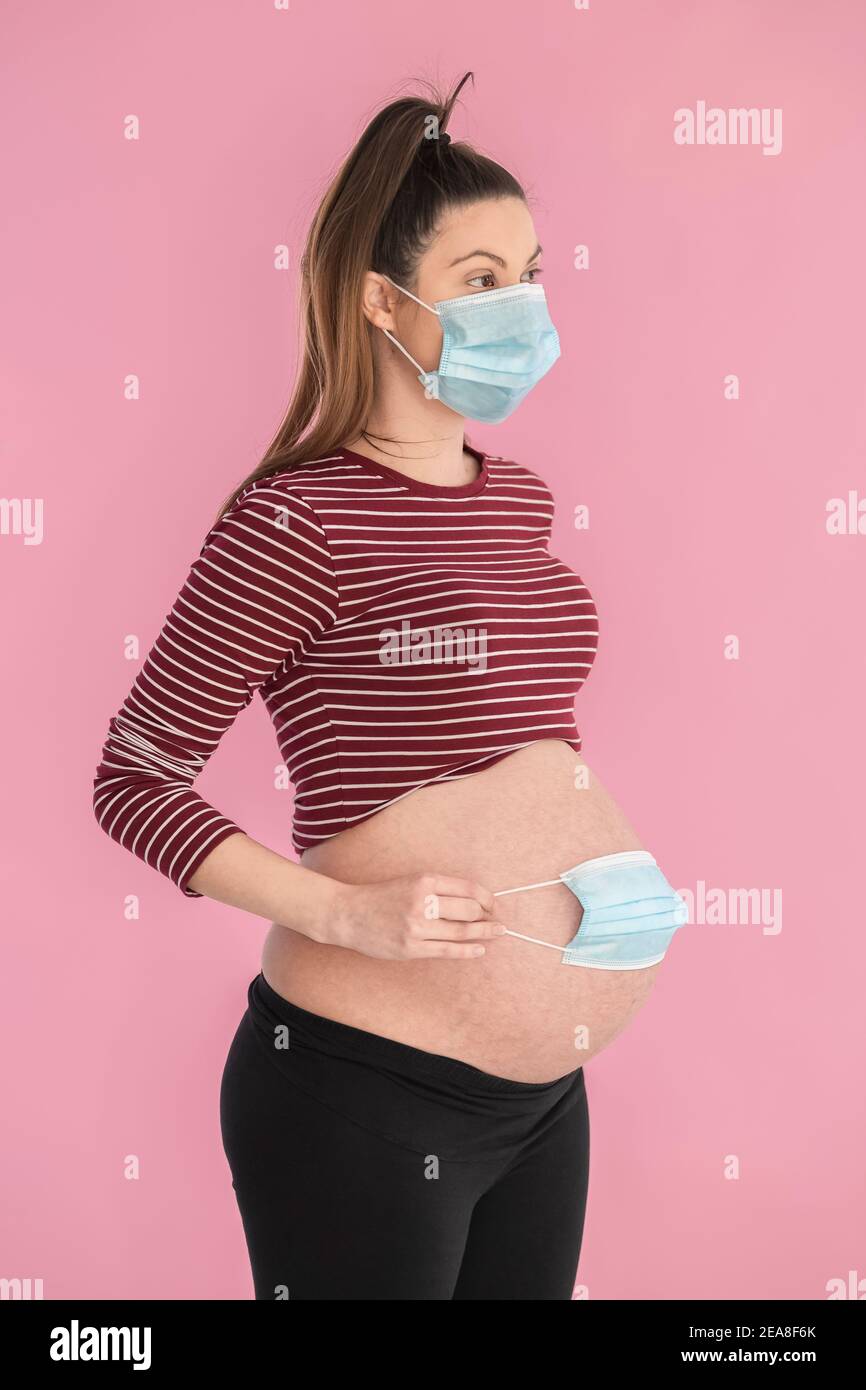 Young pregnant woman wearing a face mask holding another mask over her exposed belly on a pink
