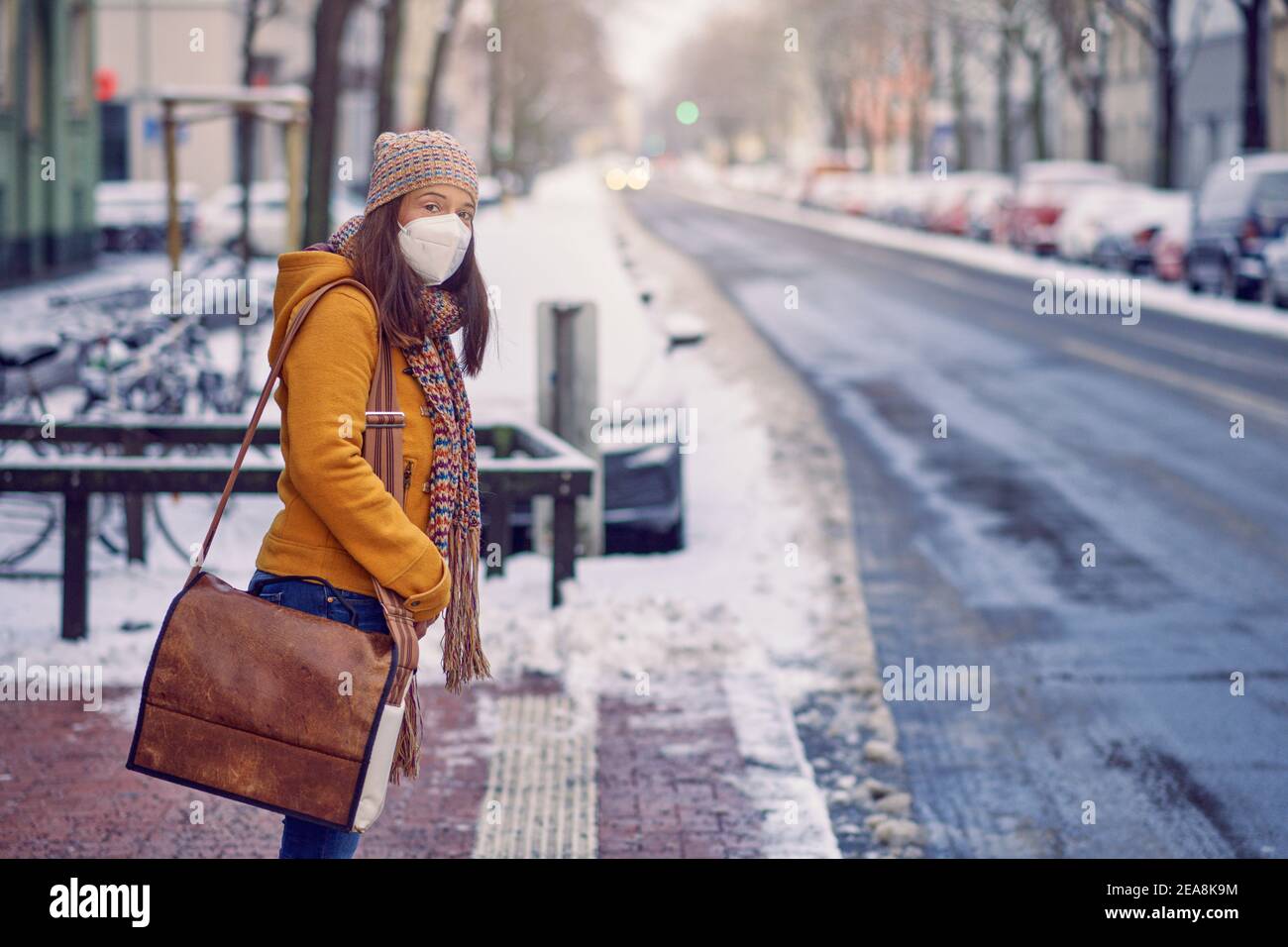Midde-aged brunette woman standing at a bus stop, wearing a protective face mask due to corona virus, waiting for her bus to bring her to work Stock Photo