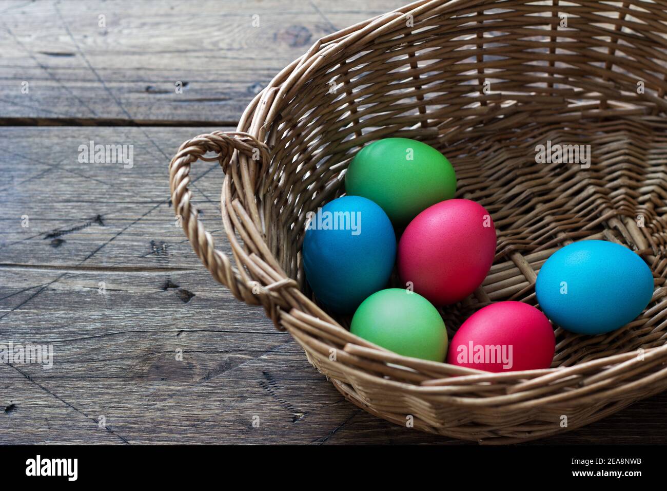 Colored easter eggs in a wicker basket on a wooden surface, space Stock Photo