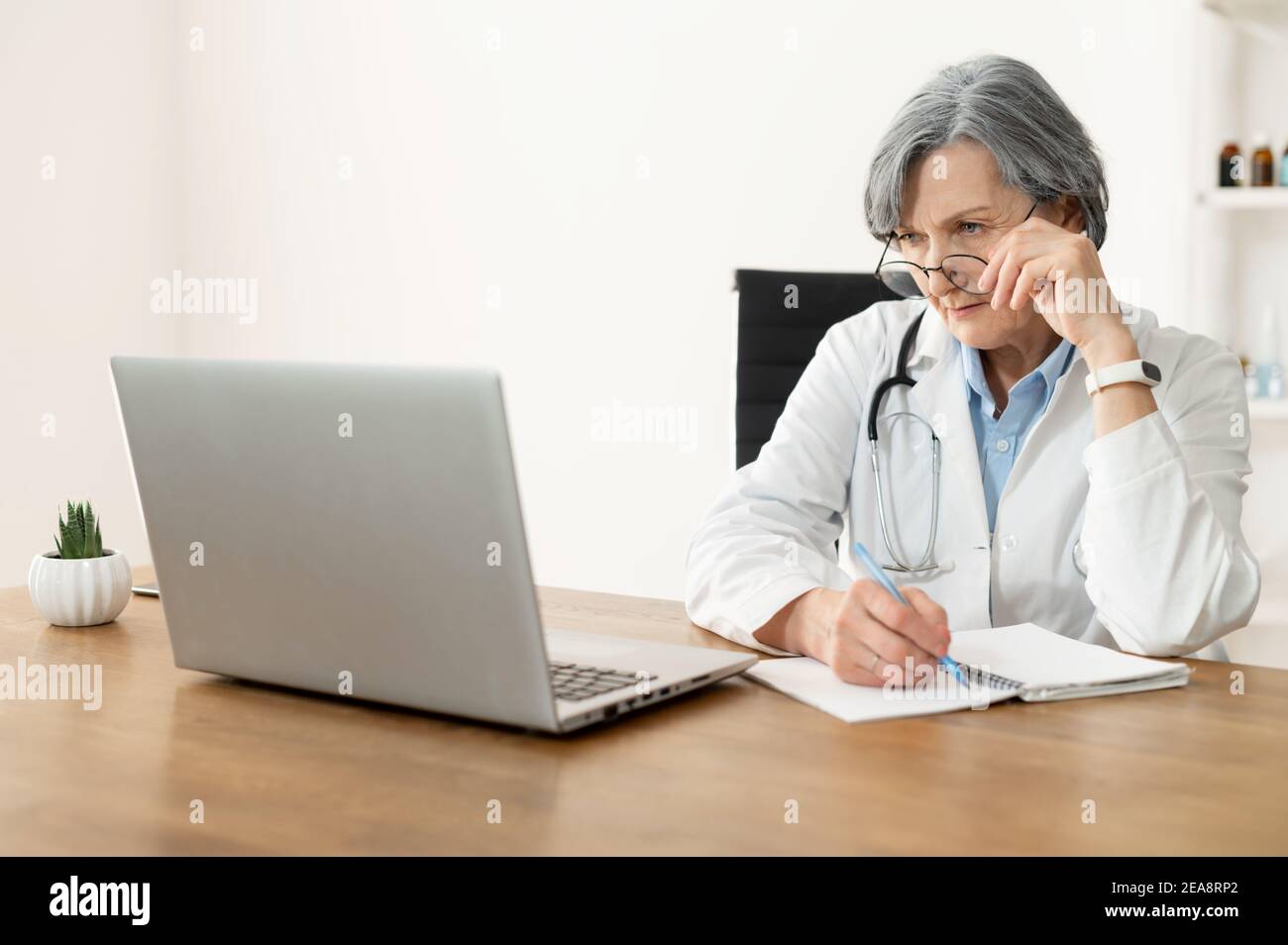 Senior old female doctor with a stethoscope in a lab coat and glasses sitting at the desk and looking at the laptop, studying online, freshening-up Stock Photo