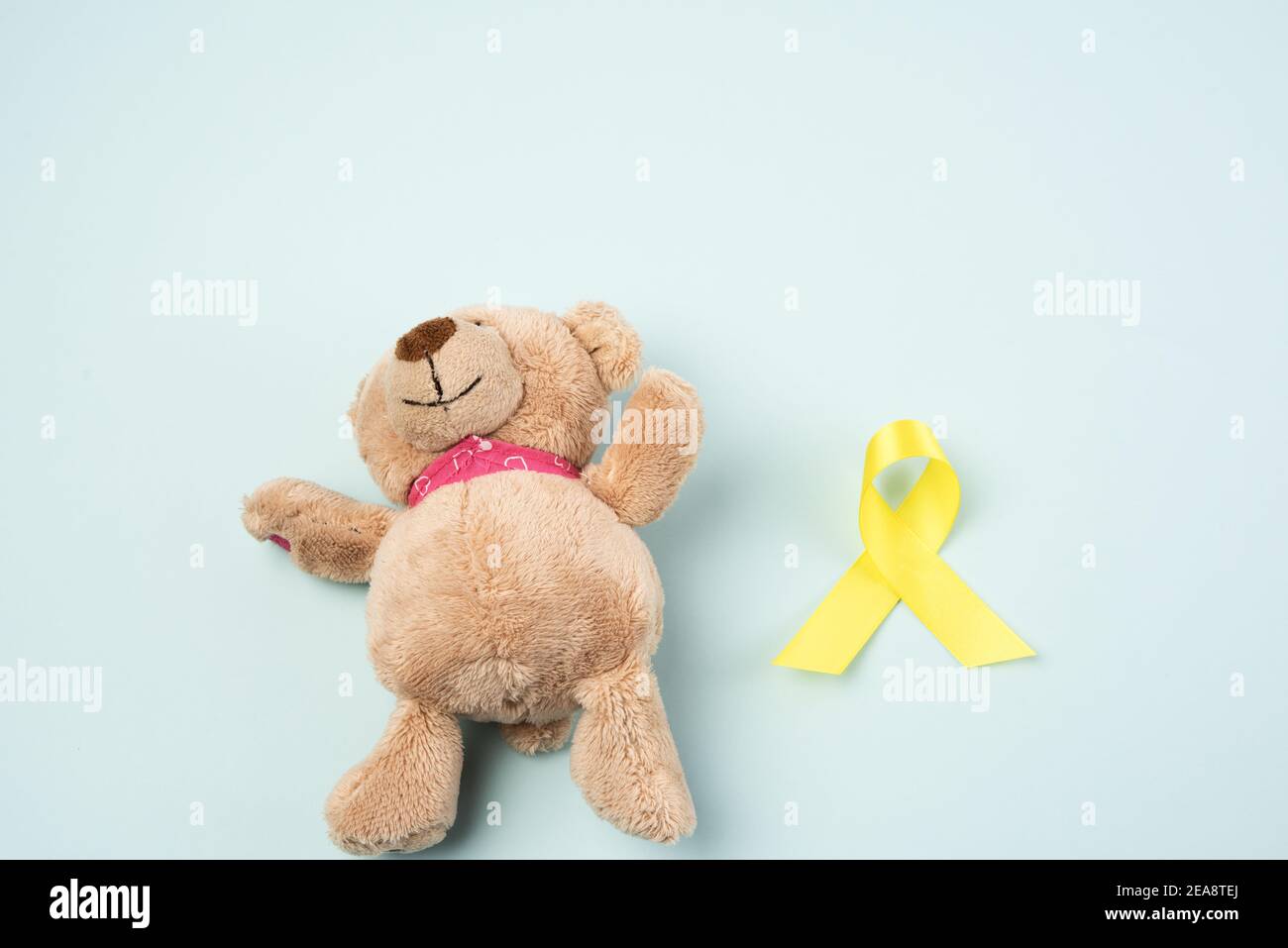 brown teddy bear holds in his paw a yellow ribbon folded in a loop on a blue background. concept of the fight against childhood cancer. problem of sui Stock Photo
