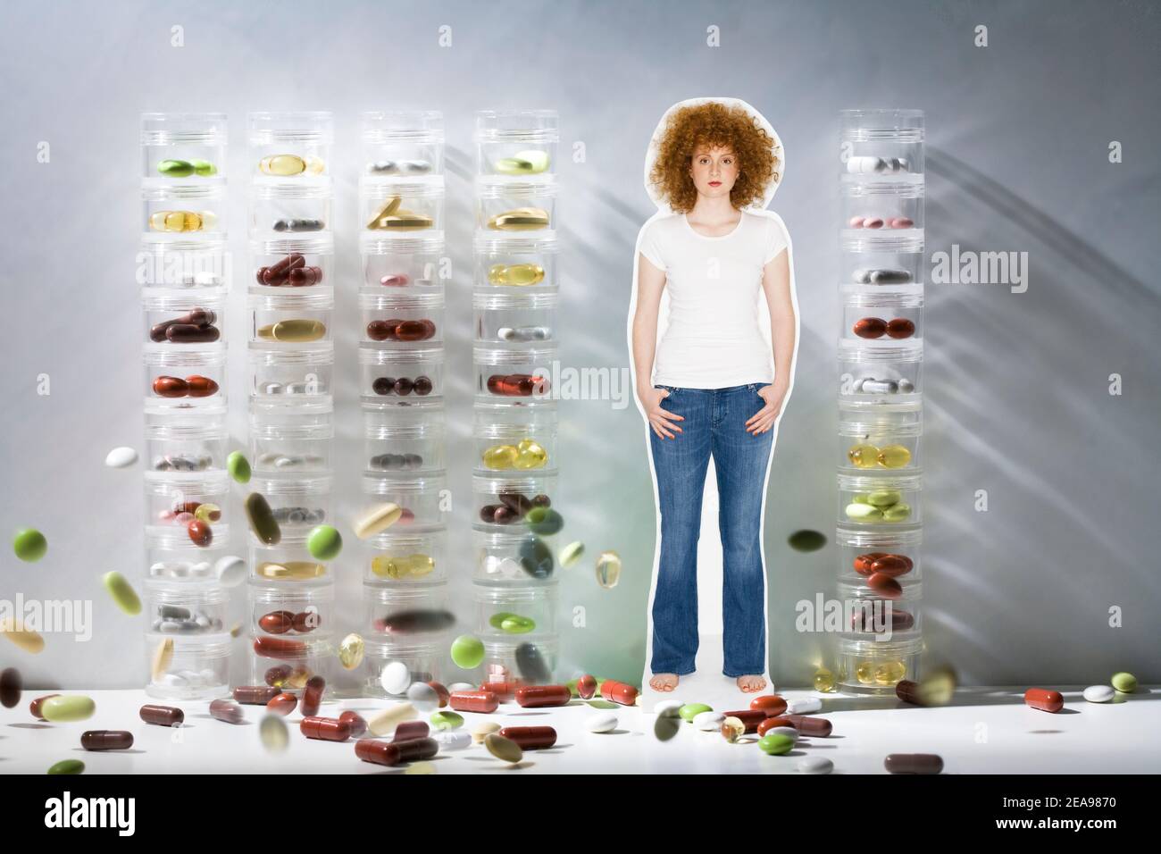 Woman in the midst of colorful tablets, in front of pill cans, white-gray background, studio, with jeans and white T-shirt, curly red hair Stock Photo