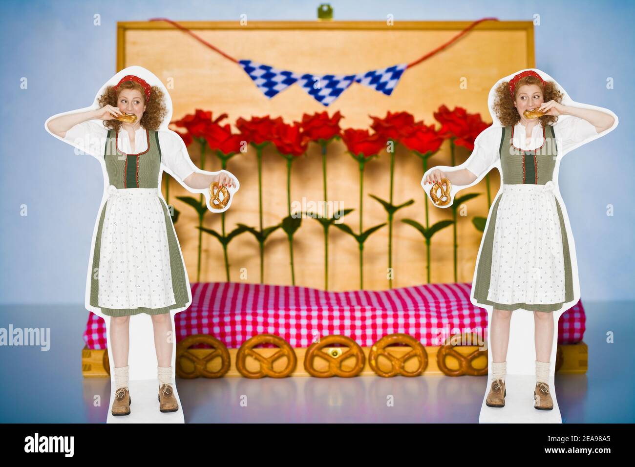 Still-life photo, 2 women in dirndls, with pretzels in their hands, biting into a pretzel, standing in front of a wooden decoration, with pretzels, Bavarian flags, plastic roses, checkered blanket, bluish background Stock Photo