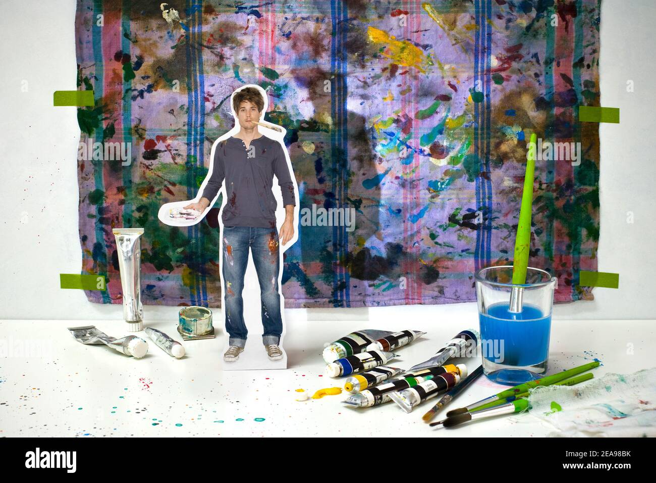 Collage photo, man in jeans and blue t-shirt, with brush in mouth, color palette in hand, stands in front of a colorful painter's background, paint tubes and water glass on a white background, colorful, blue water Stock Photo