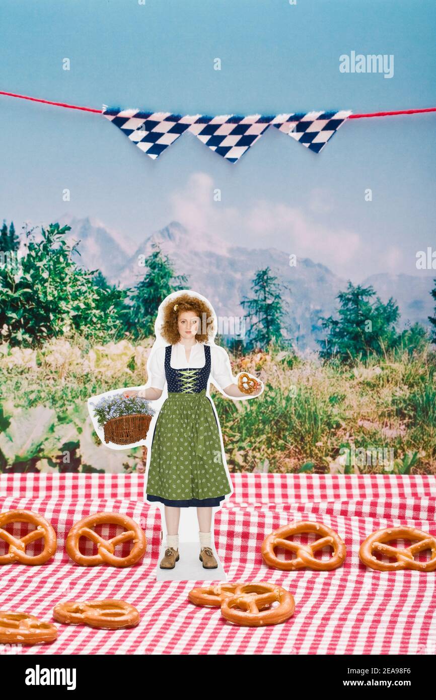 Woman in dirndl, with pretzel and flower basket in her hand, stands on a checkered tablecloth, in front of a photo of green meadow and mountains, blue and white flags hang in the background, pretzels lie on the ground, still life Stock Photo