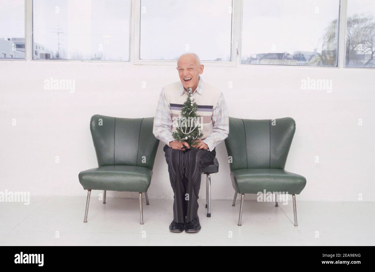 old man sits with mini Christmas tree on chair between two armchairs Stock Photo