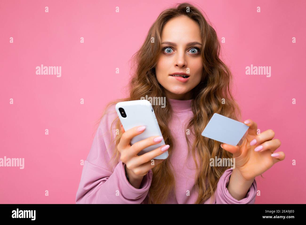 Closeup portrait photo of pretty shocked young blonde curly woman wearing pink clothes isolated over pink background using mobile phone making payment Stock Photo