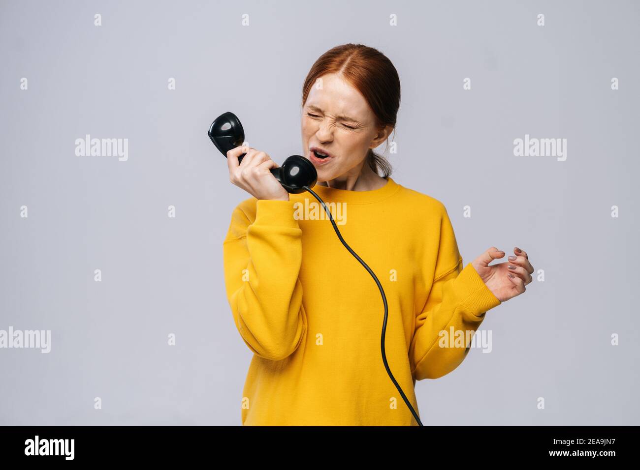 Furious angry young woman in stylish yellow sweater talking on retro phone and screaming in handset. Stock Photo