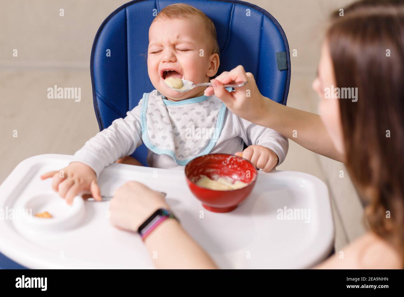 Mother tries to feed with spoon naughty infant kid refusing eating Stock Photo