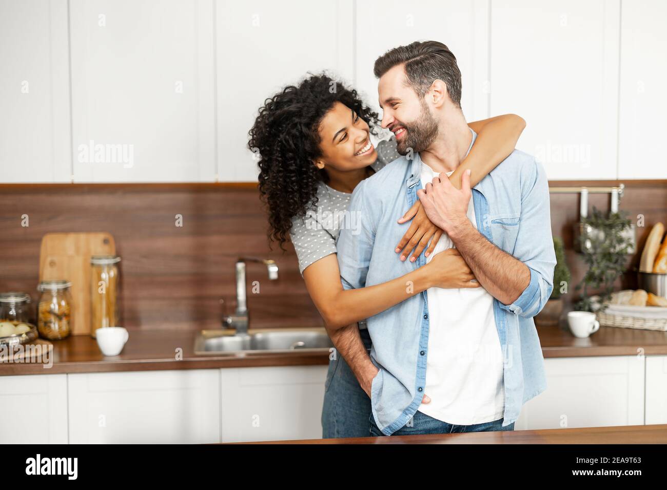 Close-up of a happy interracial couple standing in the kitchen, happy romantic owners of a new flat smiling and looking at each other, young African Stock Photo