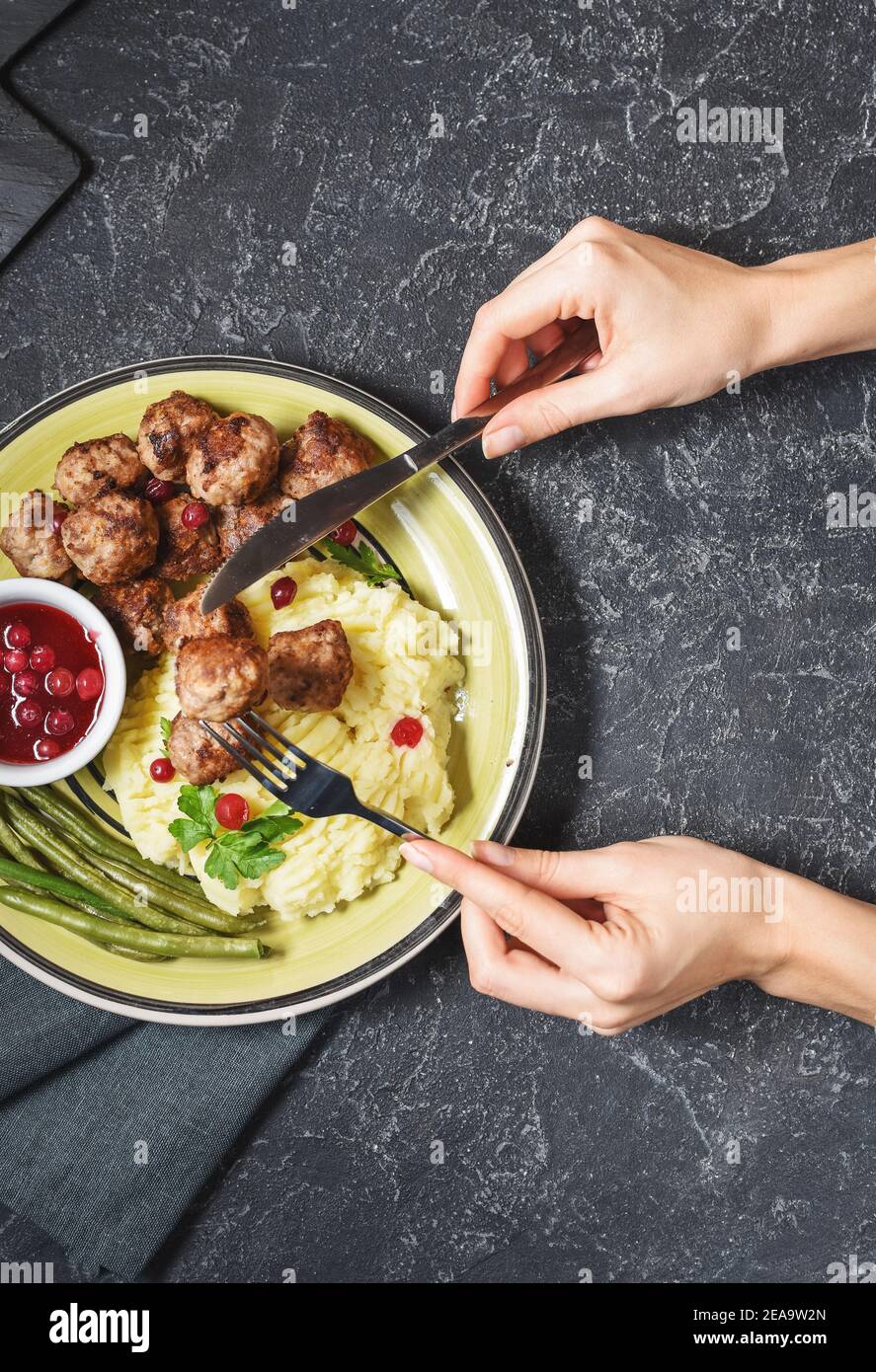 Swedish meatballs with mashed potatoes and green beans on black stone background. Female hands holds fork and knife. Top view Stock Photo