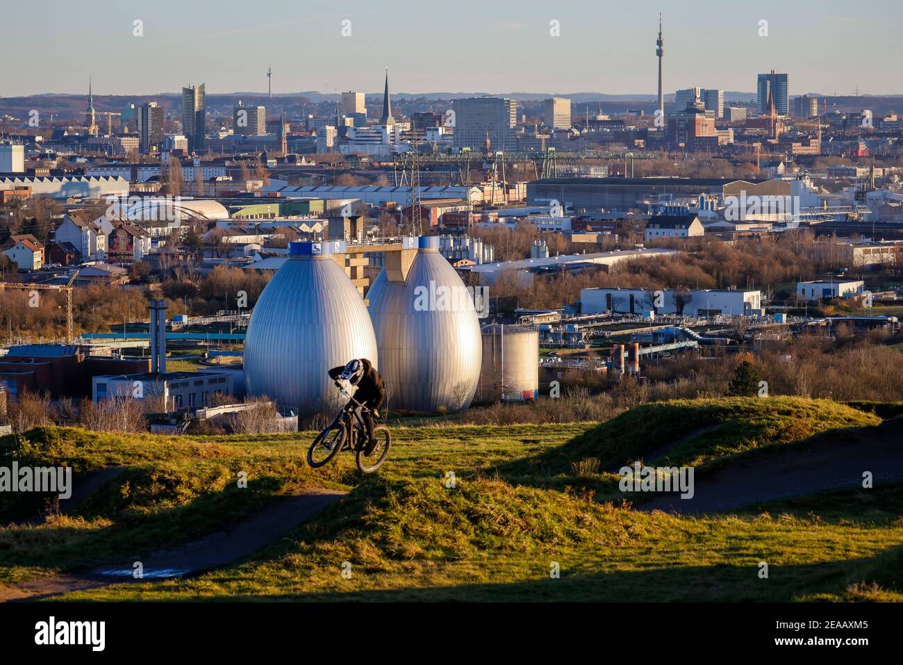Dortmund, Ruhr area, North Rhine-Westphalia, Germany - city panorama Dortmund, mountain bike riders, mountain bike arena on the Deusenberg in front of the skyline of Dortmund city center, in the back the Florian and Dortmund TV tower, in front the digestion towers of the Emscher sewage treatment plant in Dortmund Deusen, the sewage treatment plant is now receiving a 4th stage which also frees the treated wastewater from drug residues. Stock Photo