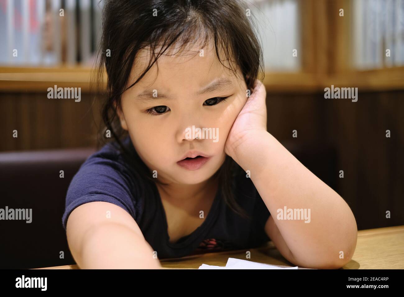 A close up of a cute young Asian girl with an expression of worry and upset on her face, resting her cheek on her hand while resting her elbow on a ta Stock Photo