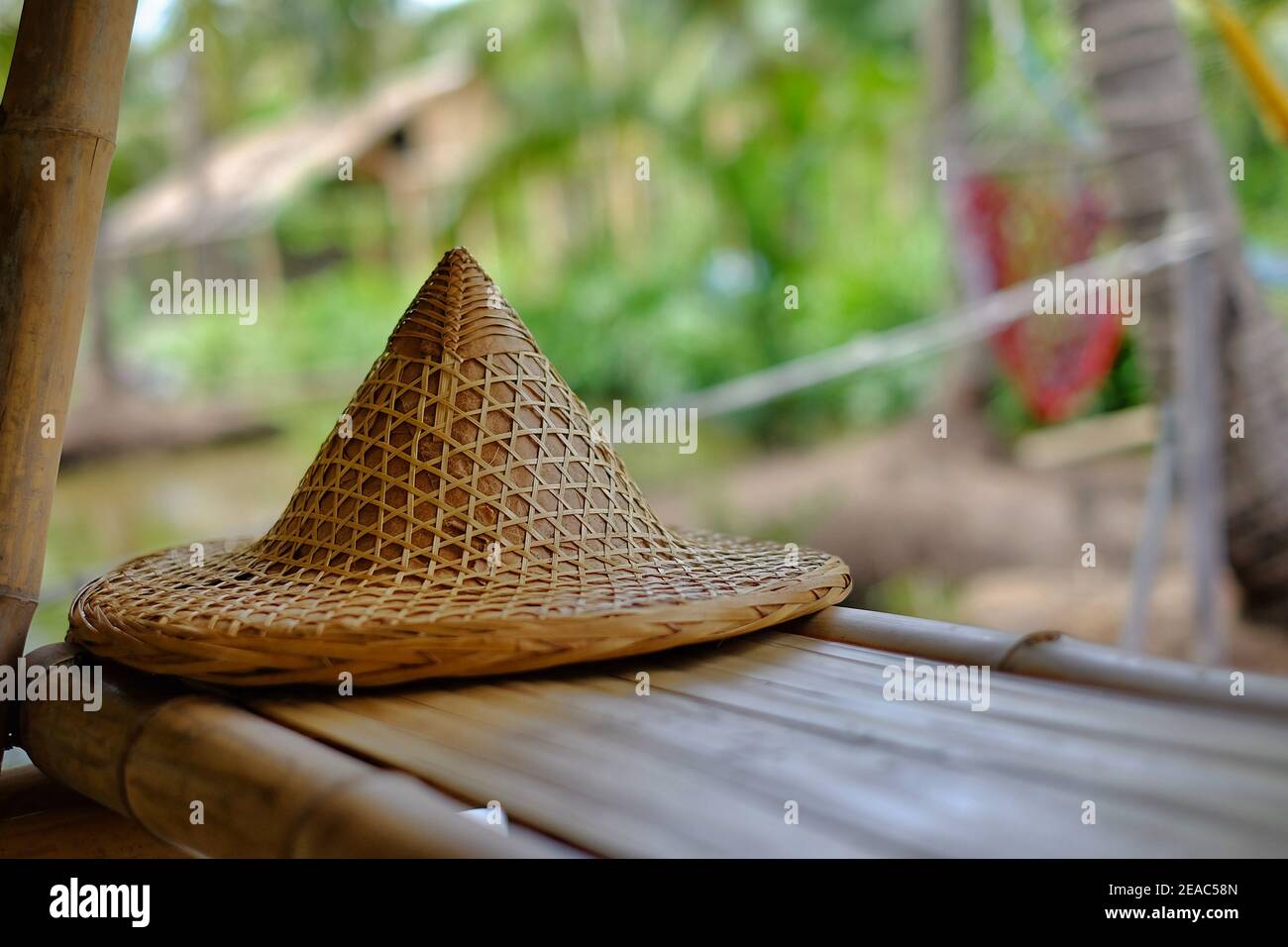 A traditional Southeast Asia farmer hat made from woven bamboo and straw is  placed on the edge of a bamboo table in a hut by a rice field in Thailand  Stock Photo -