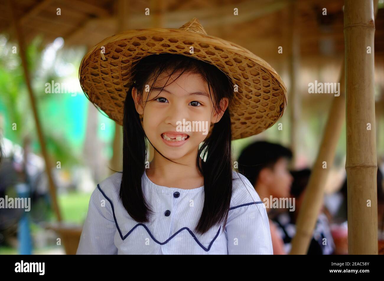 A cute young Asian girl wearing a traditional bamboo woven farmer hat at a rice farm in Thailand, feeling happy and smiling. Stock Photo