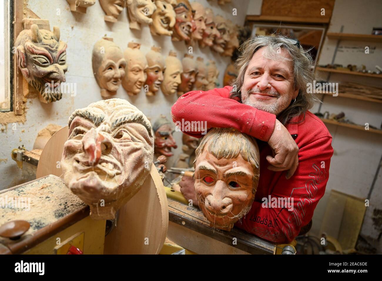 Ravensburg, Germany. 06th Feb, 2021. Jogi Weiß, mask and wood carver, leans  on a mask in his workshop while masks hang on the wall next to him, covered  in wood dust and